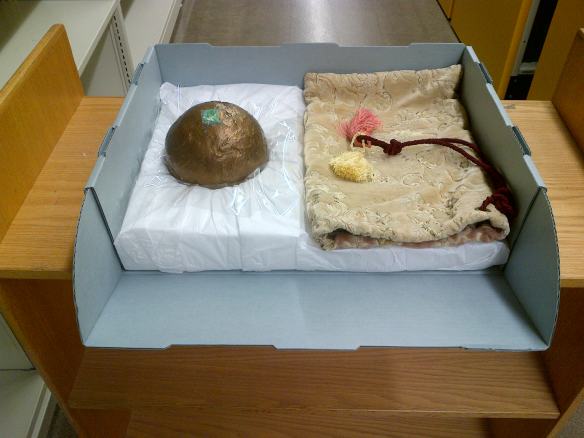 Colour photograph showing the bronze globe in a padded container with the silk brocade wrapping on the right. There is a layer of polyester film under the metal object to isolate any leaking water.