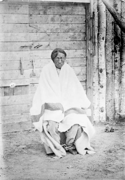 A black-and-white photograph of a man seated, wrapped in a blanket. He is looking directly at the viewer.