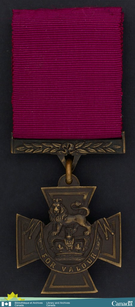 Colour photograph of a medal. Ribbon is crimson. Cross-shaped medal is bronze with a lion above a crown bearing the inscription For Valour on a scroll.