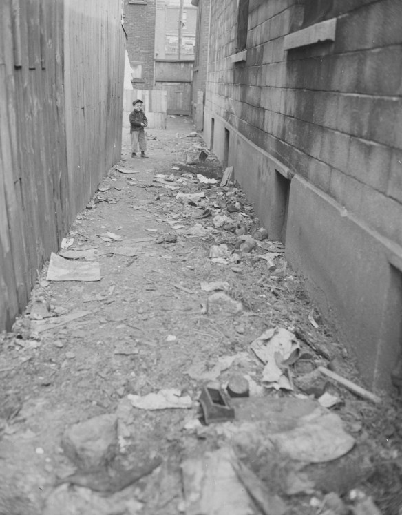 Black-and-white photograph showing a long, narrow lane strewn with garbage. A young child stands at the far end looking at the viewer.