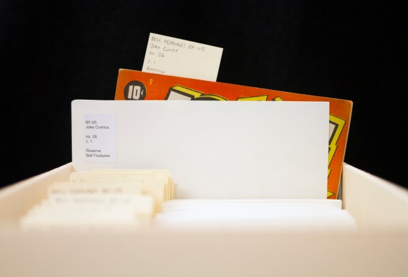 Colour photograph showing a filing box with folders with labels, one is pulled out half-way and a red comic book with the 10¢ price peeks out of the folder.