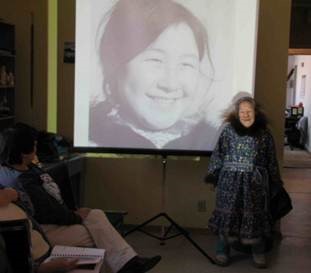 Colour photograph of an elderly Inuit woman wearing a fur-trimmed floral parka posing in front of a screen with a slide projection of her photograph when she was a young woman, taken at a community hall.