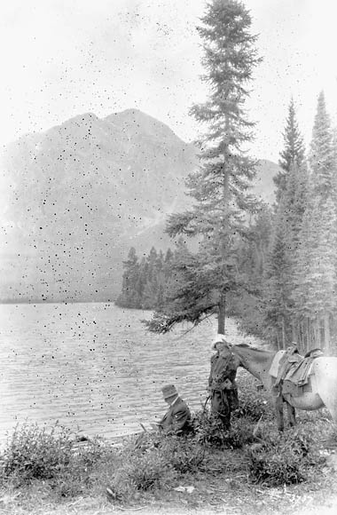 A black-and-white photograph showing a man and a woman with a horse by a lake. The man is seated and the woman is holding the lead to the horse. There are tall coniferous trees behind them.