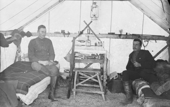 Black-and-white photograph of two men in North West Mounted Police uniform sitting on cots in a tent