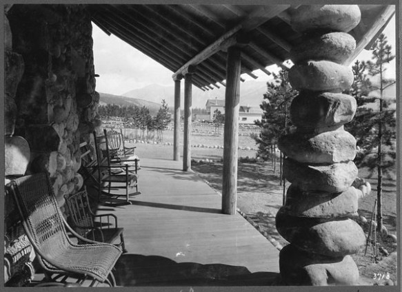 A black-and-white photograph showing a veranda with seating to take advantage of the view. The architectural style is rustic, with river stones and rough-hewn beams.
