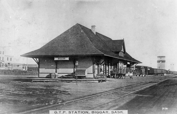 A black-and-white photograph of a train station taken from the other side of the tracks. There is a sign with the word Biggar, and there’s a note at the bottom of the photograph identifying it as G.T.P. Station, Biggar, Sask.