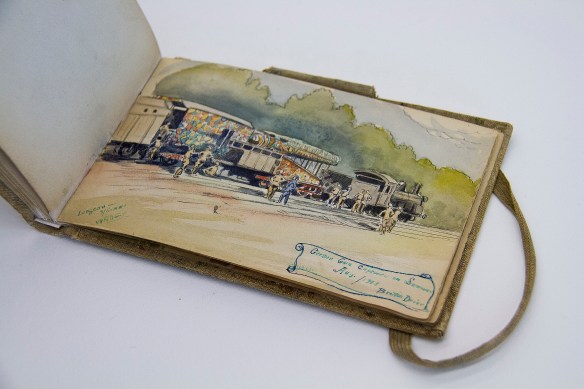 Colour photograph showing an open sketchbook with a watercolour of a train with a German naval gun on a wagon. The gun is wildly patterned with a camouflage paint. Soldiers are standing around, looking at it and talking.