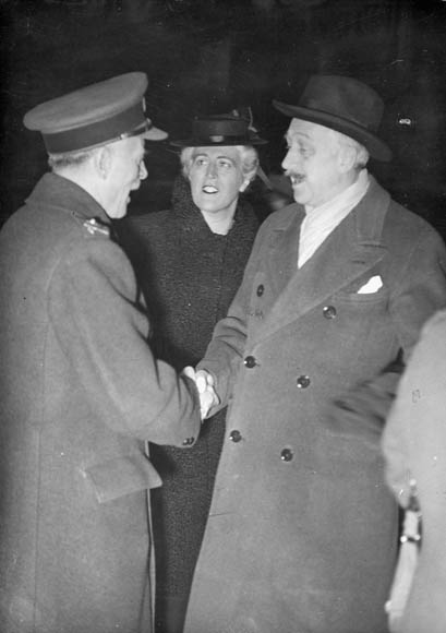 A black-and-white photograph of two men and a woman. The men are shaking hands and the woman is looking at one of the men.