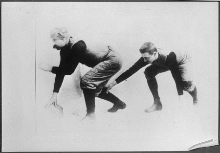 A black-and-white photograph of two rugby football players crouching, the man on the left is holding the ball, waiting to throw it to the other man behind him.