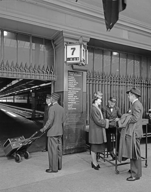 A black-and-white photograph of people in a train station. A porter, with luggage on a dolly, is facing away from the camera. Two well-dressed travellers are speaking to a ticket agent. An information board with destinations is on the wall behind the travellers announcing the train as “The Dominion” from Montréal to Vancouver. A passenger train is visible in the background.