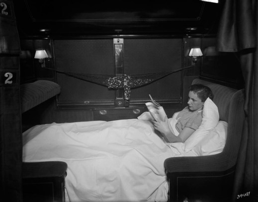 A black-and-white photograph of a woman, in profile, lying under the blankets in the lower bunk reading a newspaper.