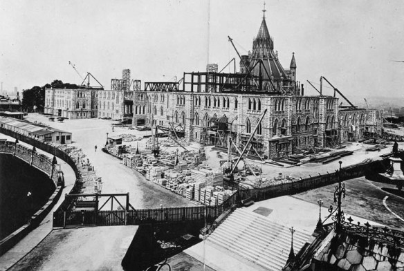 A black-and-white photograph showing the first three stories of a building with the rotunda of the Library of Parliament in the background. Cranes and construction materials surround the area.