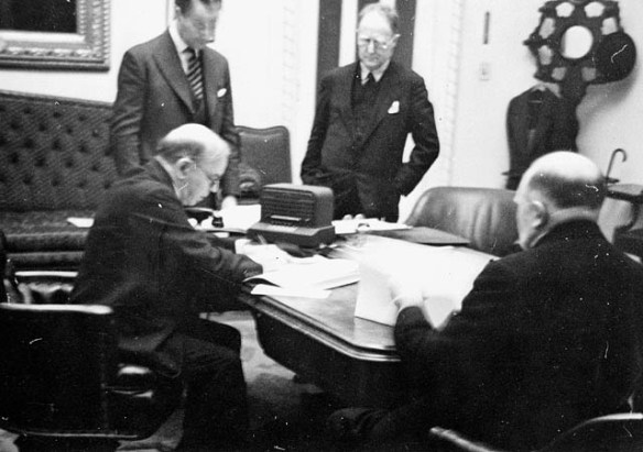 A black-and-white, slightly blurred photograph of four men in suits in an office around a table covered with documents. Two are seated and looking at papers, while the other two stand, overlooking the proceedings.
