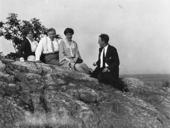 A black-and-white photograph of a group of people sitting on a high rocky ledge with blurry trees in the distant background. Everyone is looking towards the photographer except for one man who is looking at the others.