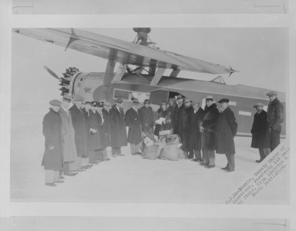 A black-and-white photograph depicting a group of men in winter coats and hats standing in front of a single-engine aircraft. The men are arranged in a semi-circle facing the camera with sacks of airmail laying on the ground.