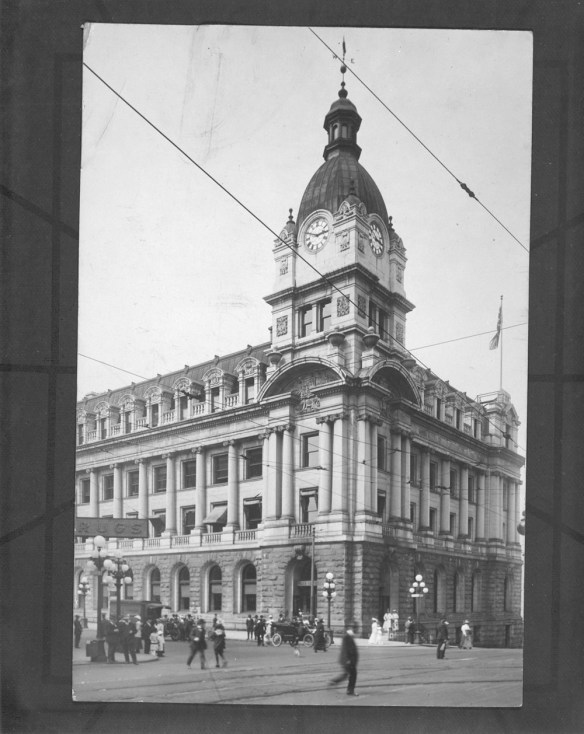 A black-and-white photograph of a very ornate building with a large clock tower in one corner. People and cars can be seen in the foreground.