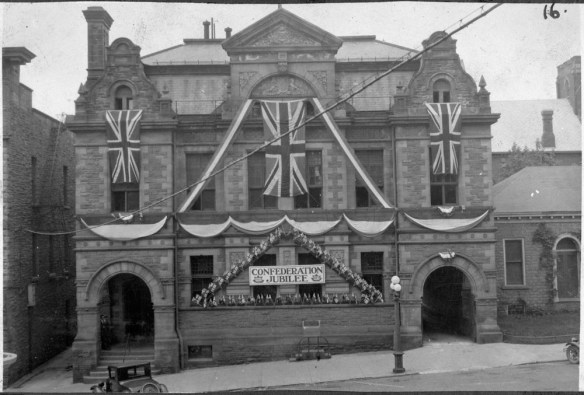 A black-and-white photograph of a building decorated with Union Jacks and a sign that reads “Confederation Jubilee.”