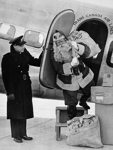A black-and-white photograph depicts a man dressed as Santa Claus exiting the side door of an aircraft carrying a sack of airmail. He is on portable steps ready to step onto the pavement. The door is held open by a man in uniform.