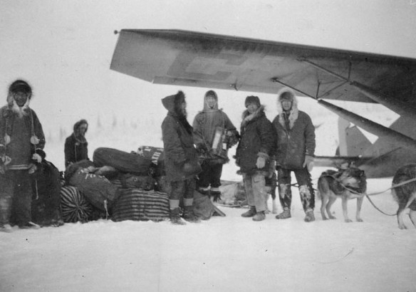 A black-and-white photograph depicting a number of men standing under the wing of an aircraft next to a dogsled loaded with sacks of airmail. Most of the men are posed, facing the camera. A number of dogs in harness are visible.