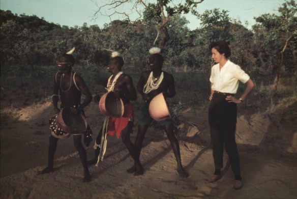 A colour photograph showing four people standing outdoors. Three are in traditional Ugandan dress while another is in western clothing.