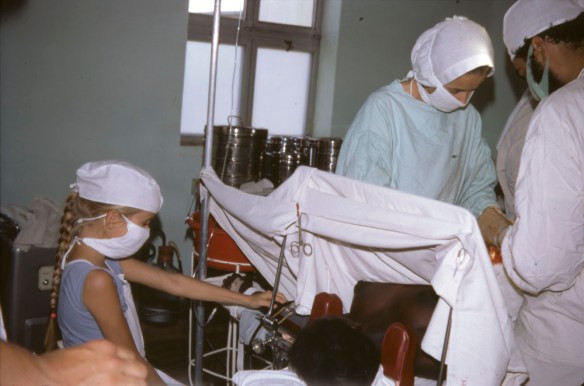 A colour photograph showing three people in an operating room. Two appear to be doctors, while the third is a child observing them.