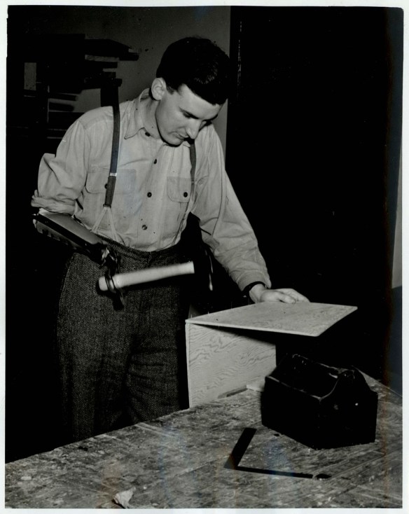 A black-and-white photograph of a man using a hammer with his prosthesis to build and assemble two pieces of wood.