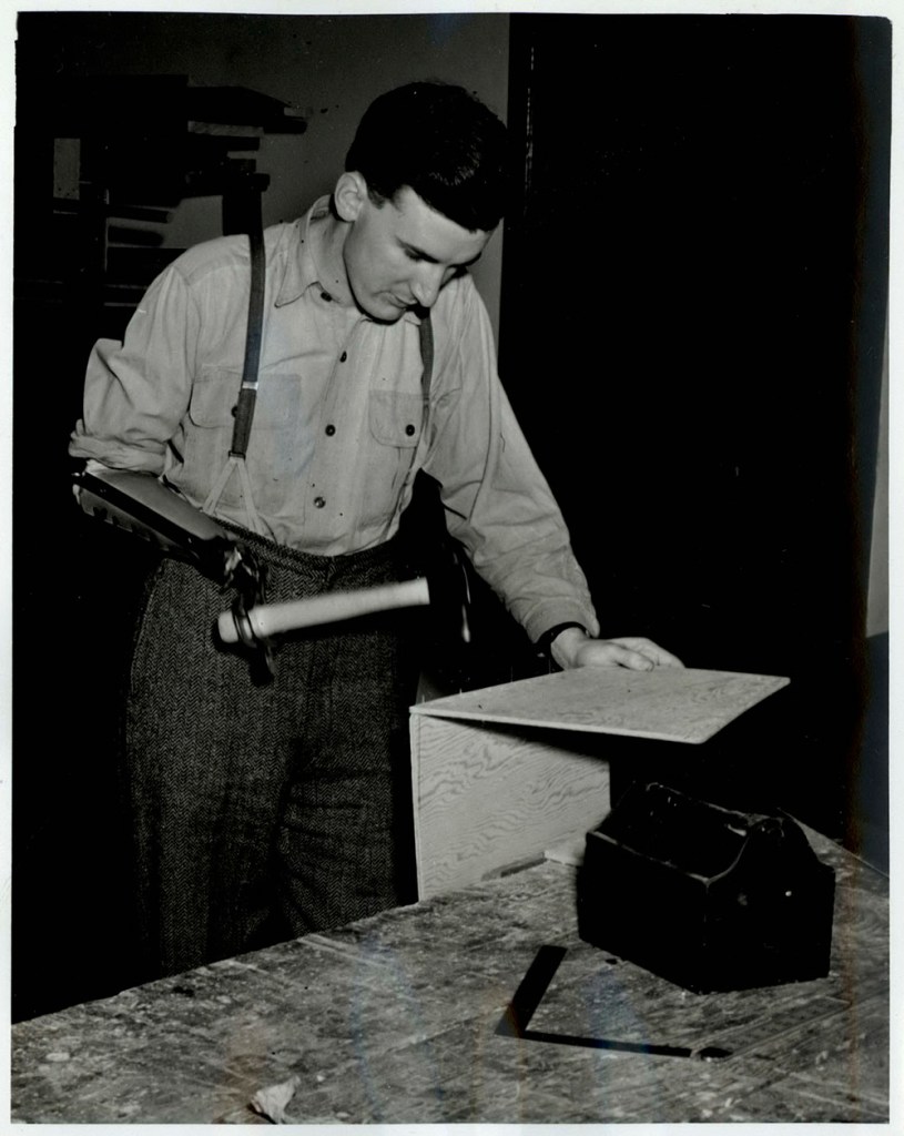 A black-and-white photograph of a man using a hammer with his prosthesis to build and assemble two pieces of wood.