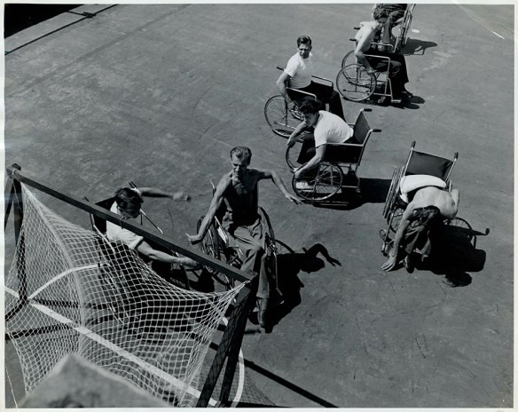 A black-and-white photograph showing seven men in wheelchairs playing a ball game around a hockey-type net. 