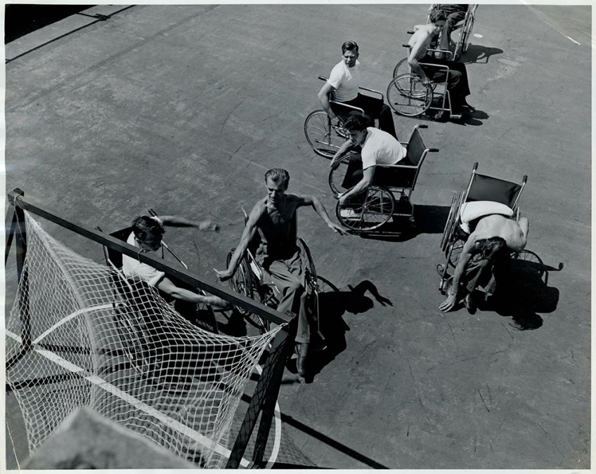 A black-and-white photograph showing seven men in wheelchairs playing a ball game around a hockey-type net.