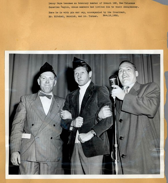 A black-and-white photograph mounted on a piece of a paper showing three men arm-in-arm around a mic. The photo is captioned.