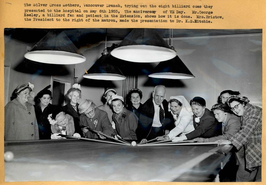 A black-and-white photograph showing a large group of women and a single man around a pool table, some of them leaning over the table and aiming at the cue ball with cues. The photo is captioned.