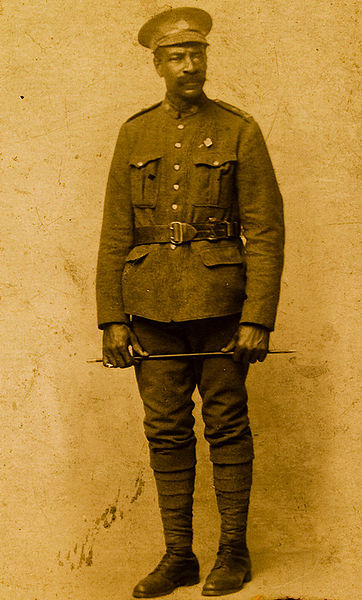 A sepia-coloured photograph of a man in uniform wearing an officer’s belt and cap holding a baton in both hands across his upper thighs.