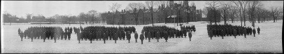 A black-and-white panoramic photograph of a battalion lined up in four groups in a snowy landscape with buildings in the distance.
