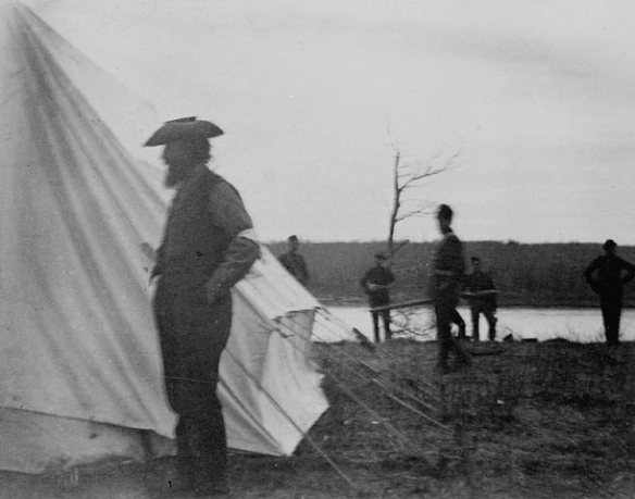 A black-and-white photograph of the side view of a man standing in front of a white canvas tent with five men standing in the background.