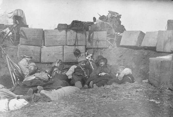 A black-and-white photograph of five men asleep on the ground behind a barricade made of earth and bundles of supplies.