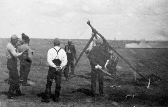 A black-and-white photograph of six men standing around an animal carcass hanging from a tripod built from branches.