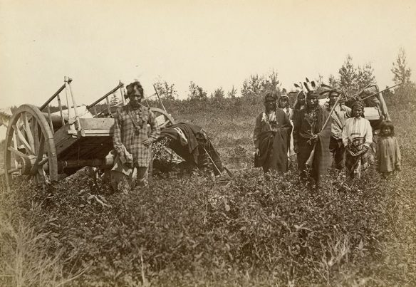 Black and white photograph of a man, on the left, wearing European clothing and standing in front of a Red River cart, and a group of First Nations men, women and children wearing First Nations-style clothing and standing in front of another Red River cart, on the right.
