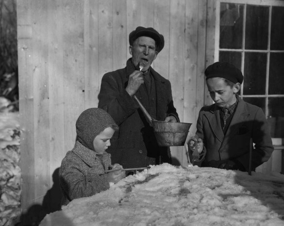 A black-and-white photograph showing three people eating maple taffy on snow.