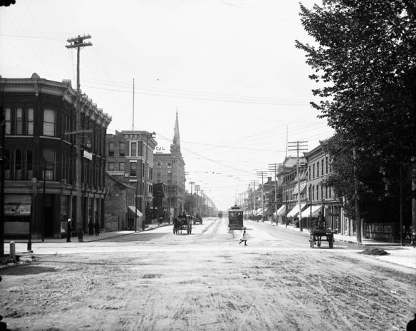 A black-and-white photograph showing a streetscape at a crossroads.