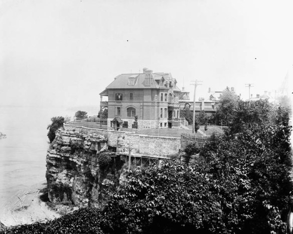 A black-and-white photograph of a large brick house perched on a cliff.