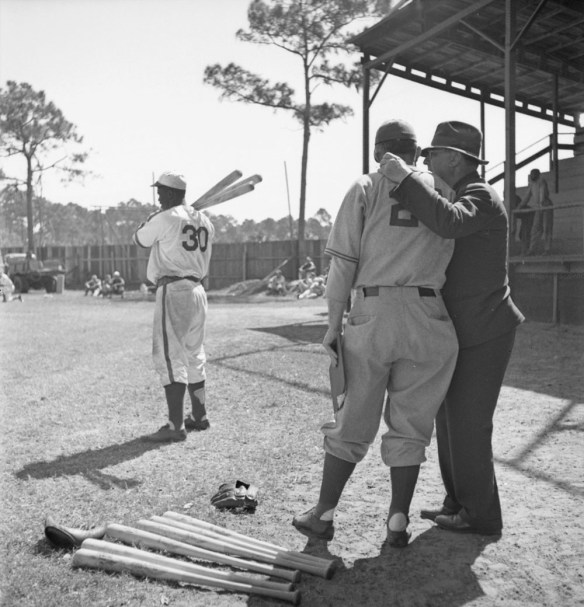 A black-and-white photograph of three men on a baseball field. Two men, one in uniform, the other in street clothes, stand in the foreground talking to each other. On the ground at their feet are two baseball gloves and a number of baseball bats. In the background, another player, in uniform, holds three baseball bats on his right shoulder. In the distance, people stand or sit on the ground and in the bleachers.