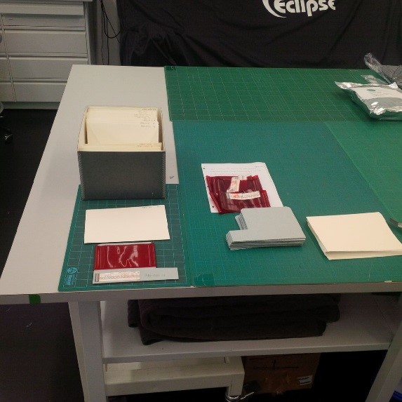 A colour photograph shows how the Dictaphone belts were received, with rusty paper clips holding the paper captions to each belt. Below the belts are the archival supplies used: blue board and envelopes. The bottom left shows the blue board inside the belt and the paper caption affixed to the bottom of the board. Above are the items placed inside envelopes and the new container that they will now be stored in.
