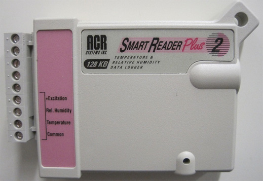 A colour photograph of a box about the size of an audio cassette that is mounted to a wall. It - records -twodifferent types of items: - Relative Humidity, Temperature-internally, along with hook-ups for external temperature and humidity sensors.