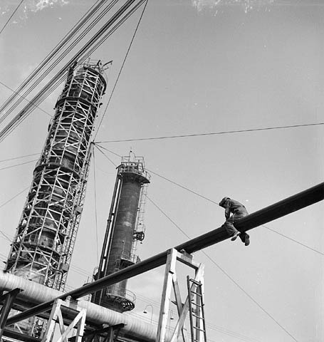 Black and white photograph taken from the ground showing, in the foreground, a welder straddling a pipe suspended in the air and, in the background, two extraction chimneys, one of which is still covered in scaffolding. 