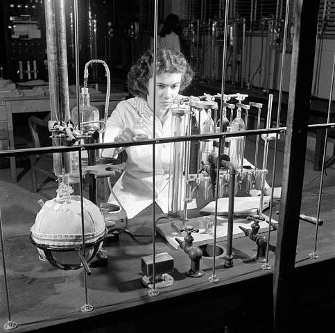 Black and white photograph showing a female laboratory technician dressed in a white lab coat, doing experiments while facing laboratory equipment. In the background, we see the back of another female laboratory technician as well as other equipment.