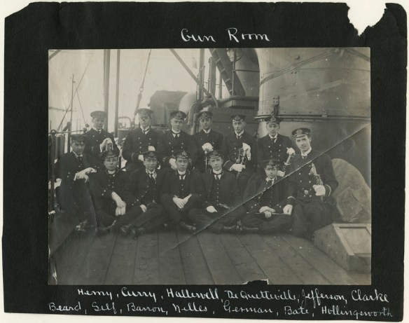 A black-and-white photograph showing a group of men sitting or standing on the deck of a ship. The photo is mounted on a black background with the words “Gun Room” written at the top and a list of names at the bottom.