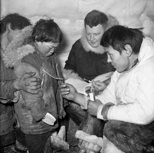A black-and-white photograph taken inside an igloo of two men reading a disc number attached to a boy’s parka.