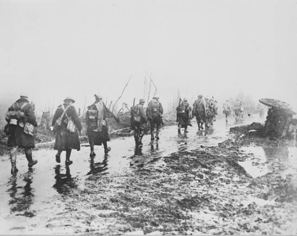 A black-and-white photograph of soldiers walking away from the photographer down a narrow muddy track.
