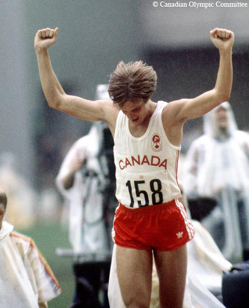 Colour photograph of a man, dressed in shorts and sleeveless t-shirt, standing with his arms raised in the air. In the background are people dressed in rain gear.