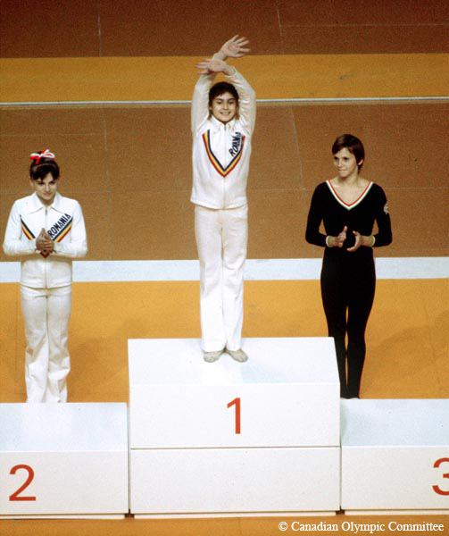 A colour photograph depicts a young woman standing on the podium waving to the crowd. She is wearing a white track suit with “Romania” written on it. Behind her on the floor are two other young women.